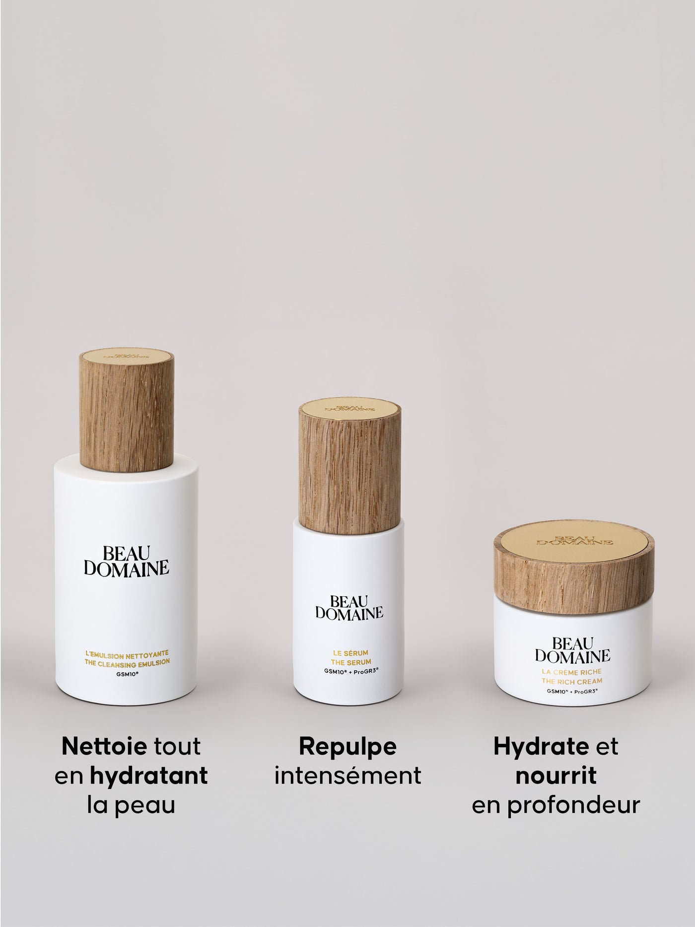 Détail des trois produits, contenant à la fois l'émulsion nettoyante, le sérum et la crème riche sur un fond beige et épuré et avec le titre des produits. Ils ont leurs capuchons en bois.